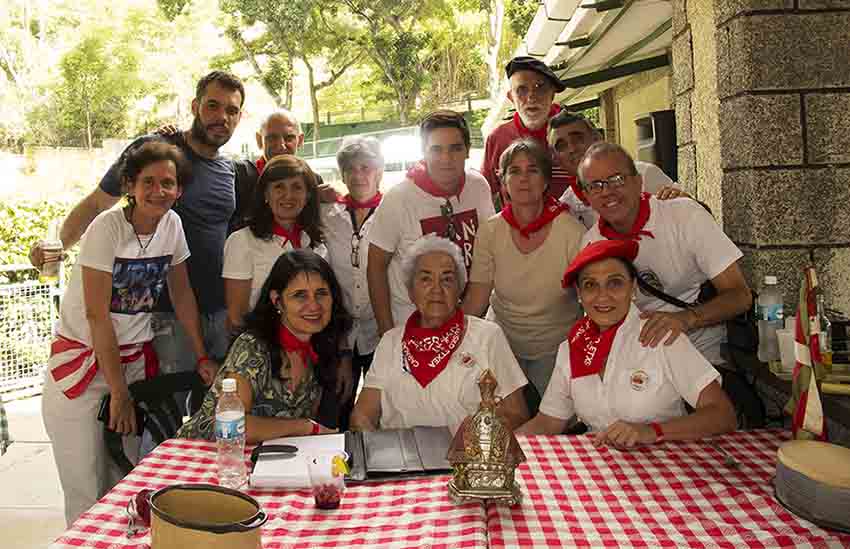 Epaile zorrotzak mahaian eserita eta partaide pintxogileak atzean San Fermin Pintxo Lehiaketaren irudian